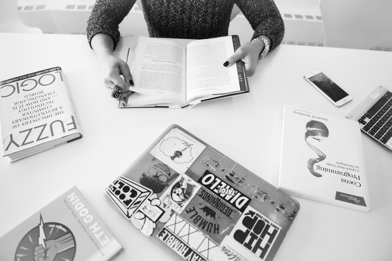 team-01 Person reading amidst books and technology on a table in a monochrome setting.