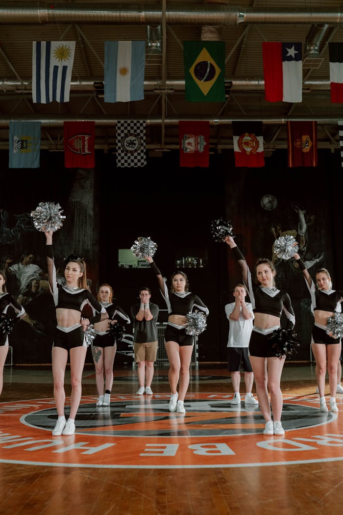 Cheerleaders in uniform performing with pom-poms inside a gymnasium under international flags.