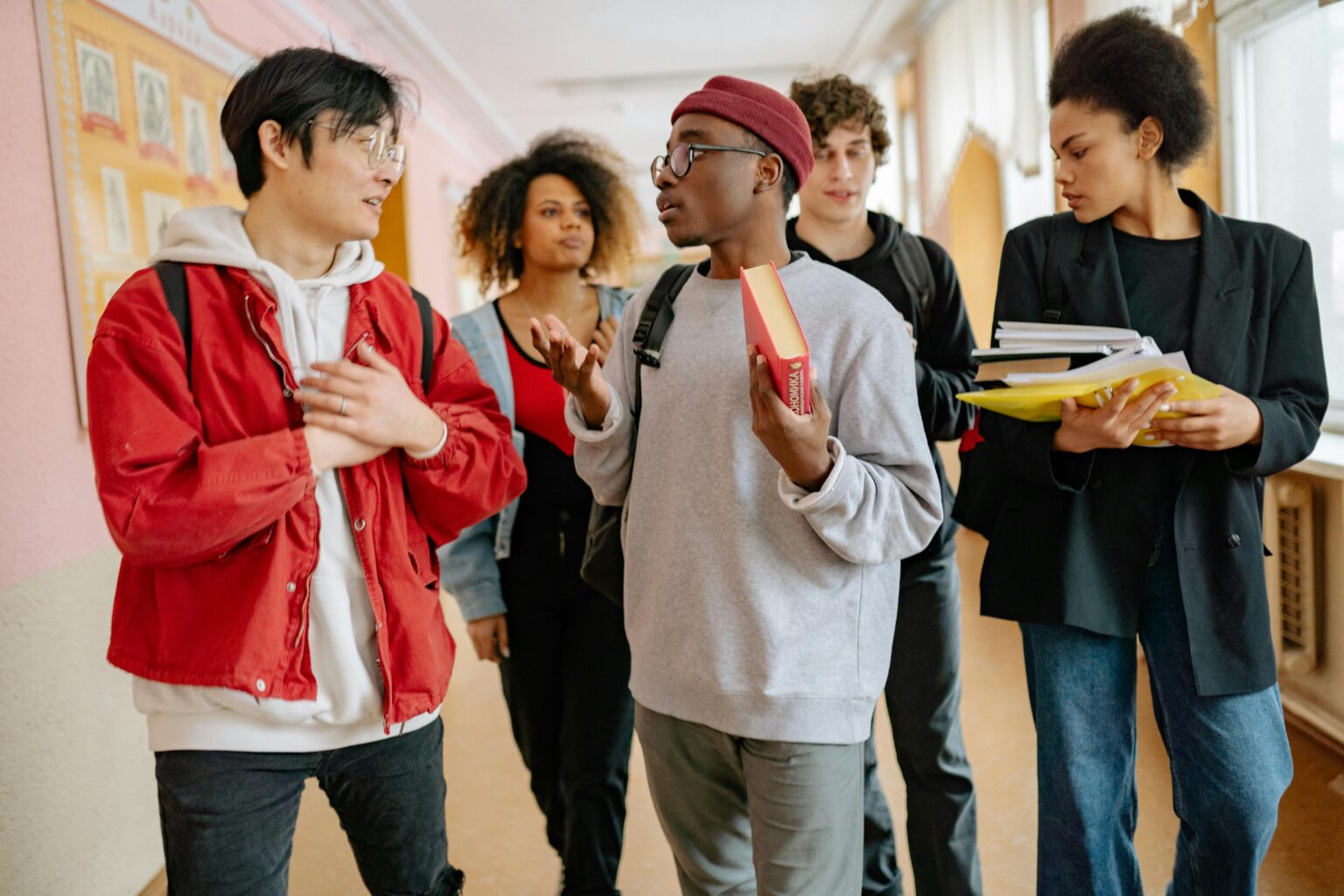 A group of diverse students walking and talking in a school corridor, carrying books and folders.